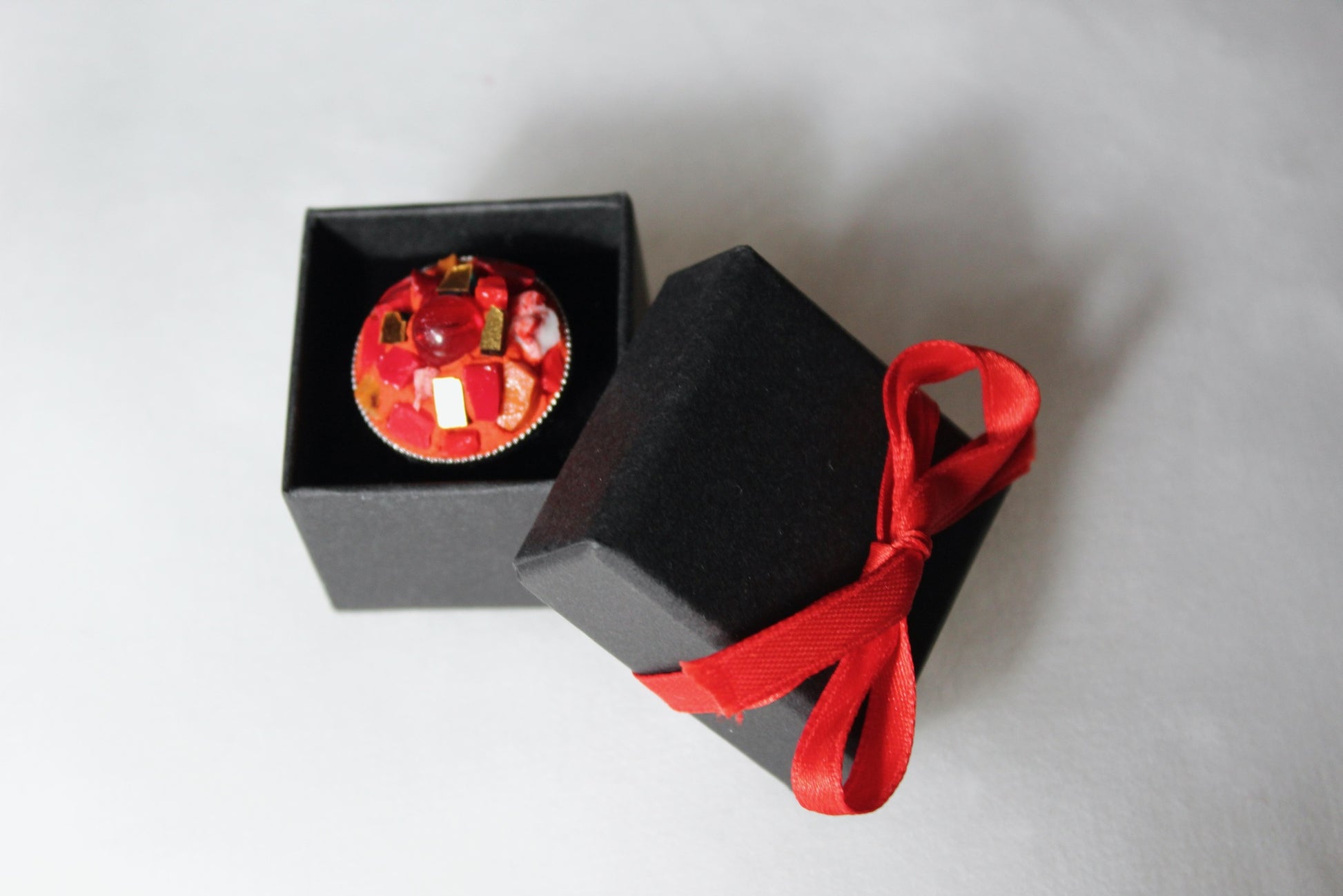 Anello in micromosaico rosso e oro con perla su sfondo bianco nella scatolina nera con fiocco rosso