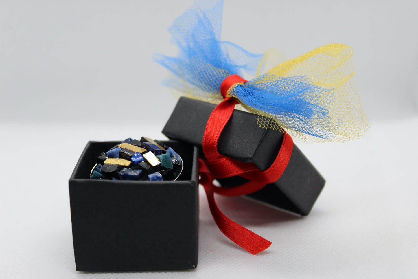 Spilla tonda blu in micromosaico con oro nella scatolina nera con fiocco rosso su sfondo bianco 